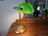 Front angle view of bankers desk lamp showing green glass shade, brass stand and base on wood surface with mirror background.