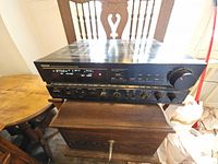 Front angle photo of black Denon Precision Audio Component AM/FM Stereo Receiver sitting on a wooden chair. Shows front control panel, knobs, and display.