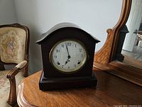 Photo of a dark mahogany desktop clock with arched top and white face with black numbers, sitting on polished wooden furniture with a tapestry chair and mirror in the background.
