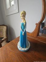 Full view of Elsa figurine standing on wooden surface, next to a mirror with a window in background.