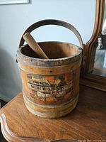 Large antique wooden bucket with metal handle containing a large wooden pestle. The bucket shows a worn business sticker and natural wood wear.