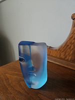 Front and side views of the blue and frosted glass face sculpture placed on a wooden surface with a mirror in the background.