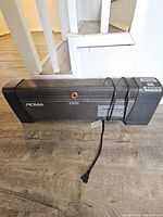 Photo showing the full front view of the black NOMA space heater model 8006 placed on wood flooring with power cord wrapped around the heater body.