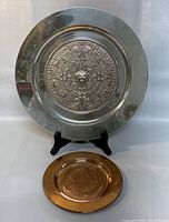 Two round metal wall plates on a stand; vintage Mexican Alpaca plates with embossed Aztec calendar design, silver and copper toned