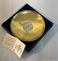 Round gold-toned decorative plate with central silver bee medallion and accompanying information card in display box.