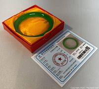 Natural green jade bangle displayed in red box with yellow cushion and certification card visible.