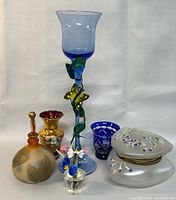 Full view of lot showing all glass pieces: Victorian satin glass trinket box, tall blue vase with butterfly, amber bottle with stopper, ruby red miniature vase, blue cut to clear miniature cup, and two small glass flower clusters.