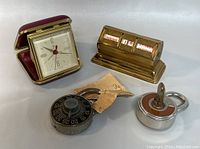 Overview of all four items in the lot including travel alarm clock, brass rolling calendar, two vintage locks (combination padlock and key lock).