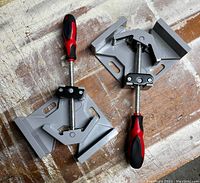 Two corner clamps placed on a wooden surface, visible grey metal body with screw and red-black handles.