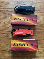 Two Premier Edge survival folding pocket knives, one black and one orange, shown alongside their branded boxes on a wooden surface.