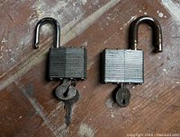 Two used padlocks side by side on a wooden surface. Both have ribbed metal bodies and keys attached. One padlock is unlocked with rust on the shackle, the other is locked.