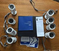 Eight white ANNKE cameras, user manual, quick installation guide, and security stickers arranged on a wooden surface.