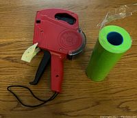 Red MX 5500 price tag gun with wrist strap, with a roll of green number tags beside it on wooden surface.