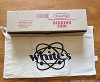 Lesche branded box with digging tool and White's canvas pouch beneath it showing company logo.