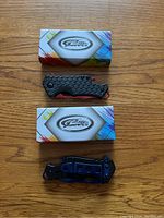 Both Rite Edge pocket knives side by side next to their boxes on a wooden surface. Red and blue blades visible folded in black textured handles.