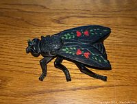 Front angled view of a black cast iron fly shaped match safe with red and green painted floral accents on wings, showing its detailed legs and textured body.