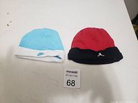 Two baby hats placed side by side on a white surface. Left hat is light blue and white with Nike logo; right hat is red and black with Michael Jordan Jumpman logo.