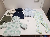 Five baby outfits laid out on a bed: white and olive green romper, navy blue vest, two footed onesies in pastel blue and yellow floral patterns, and a fleece dinosaur hooded jacket.