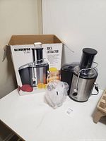 Juice extractor blender with black and stainless steel body alongside original product box and a plastic-wrapped clear juice container.