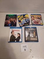 Photo of five Blu-ray DVD cases including Sex and the City, Police Academy, Wedding Crashers, The Proposal, and There Will Be Blood, arranged on a flat surface.