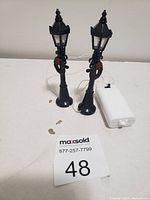 Two black street lamp style Christmas lights with red wreath decorations alongside a white battery pack on a white surface.