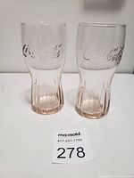 Two clear Coca-Cola glasses with embossed logo and amber tint near bases, standing side by side on white surface.