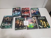 Photo of seven DVD box sets from the CSI TV franchise arranged on a white surface. Titles include CSI: Crime Scene Investigation, CSI: Miami, and CSI: NY.