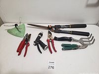 Photo of seven garden tools arranged on a white surface including shears, pruners, a hoe, a hand cultivator, and hose spray nozzle.