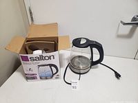Photo of the Salton electric hot water kettle next to its original box, showing the glass body, black handle, and power cord on a white surface.