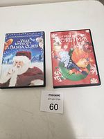 Two DVD cases on a white surface with MaxSold lot tag number 60 visible. The DVDs are "The Year Without Santa Claus" and "A Miser Brothers Christmas."