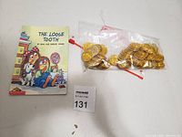 Photo of children's book 'The Loose Tooth' and two bags of gold-colored play coins shown together on a flat surface with a lot number tag.