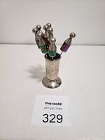 Set of six olive forks with colorful fabric handles and silver tips arranged standing in a silver-tone metal stand, photographed from front