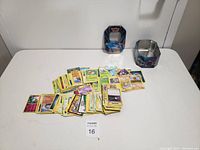 Wide view showing a pile of Pokemon cards with two metal tins placed behind them on a white surface