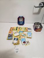 Photo showing a pile of assorted Pokémon cards including holographic cards and a hexagonal metal storage tin.