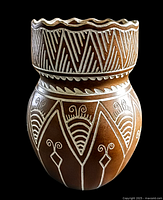 Front view of brown pottery vessel with white carved geometric and linear patterns and scalloped rim.