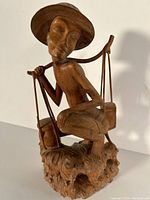 Front and full view of wood carving showing figure wearing a broad hat, sitting on a detailed base and carrying buckets on a pole over shoulders.