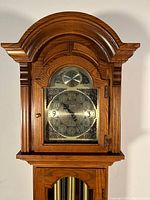 Front view of wooden grandfather clock showing arched top and brass clock face with numerals and decorative corners.