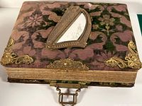Photo album top and spine showing velvet floral cover, brass corner protectors, decorative front frame with partial mirror, and clasp closure