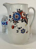 Front side view of the white ceramic pitcher with colorful floral painting and handle, alongside a water bottle for scale.