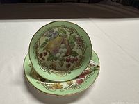 Teacup placed on the matching saucer viewed from above showing fruit design and gold detailing on green background.