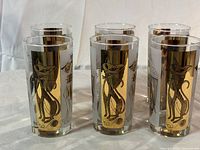Six identical clear cylindrical glasses with gold metallic panels featuring black outlined drawings of cats, dogs, parrots, and fish arranged on a white cloth surface.