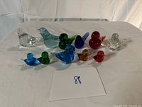 Front angle view of all 11 glass bird paperweights arranged in two rows on a white surface with neutral background.