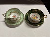Front view of both teacup and saucer sets showing floral and gold designs, one pale green and one dark green.