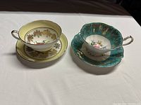 Two pairs of teacups and saucers featuring floral and gold accents, one yellow and one turquoise set.