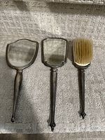 Front view of the shield-shaped hand mirror, rectangular hand mirror, and hairbrush with natural bristles, all with sterling silver frames and handles, showing surface wear and patina.