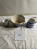 Three ceramic pieces placed on a white surface including a small green striped cup, blue and white striped bowl with spout, and small blue striped pitcher.