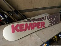 Bottom side of the snowboard showing the white base with pink Kemper logo and starburst design, minor marks visible on surface.