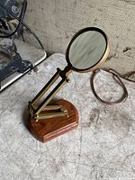 Magnifying glass showing brass frame, adjustable arm, and wooden base on a textured surface with other objects nearby.