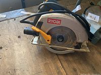 Photo showing a Ryobi circular saw on a wooden workbench, blade covered with a metal guard, power cord attached and coiled around unit, and yellow adjustment lever visible.