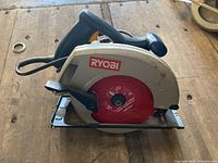 Ryobi circular saw placed on a wooden surface showing the power cord, handle, blade guard with Ryobi branding, and metal base.